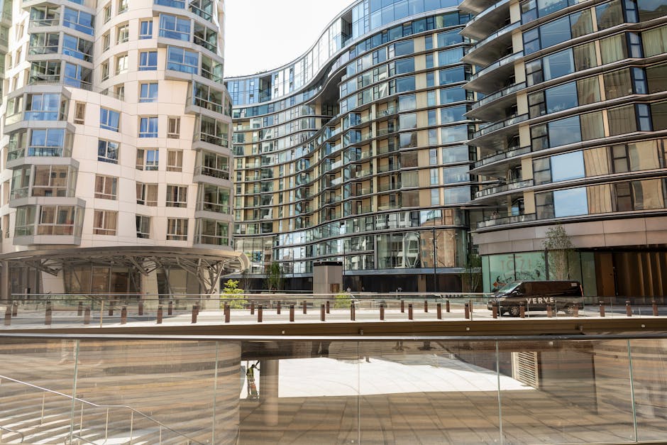 An exterior view of a modern residential building with a curved glass facade and multiple balconies, situated in an urban environment with a paved pedestrian area in front. The foreground features a glass railing, and a black van is parked near the building's entrance. The scene is well-lit with natural daylight, and the building's contemporary architecture is characterized by reflective glass surfaces and metallic accents. This setting illustrates a typical location for house removals or furniture transport, where moving companies like elephantandcastlemanandvan.com assist in home relocation and packing and moving services for apartments around Elephant and Castle.