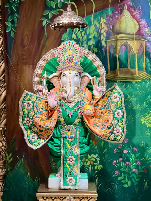 A detailed, colorful statue of the Hindu deity Ganesha, featuring an elephant head with large ears and a curved trunk, dressed in ornate orange and green clothing with floral patterns. The statue is adorned with jewelry and a crown decorated with vibrant jewels, and is positioned on a white pedestal inside a decorated indoor space. Behind it, there is a vivid painted wall mural depicting lush green foliage and a small ornate fountain with a golden structure. A decorative metal hanging lampshade is suspended above the statue. The setting suggests a religious or cultural display, with careful attention to intricate decorations, bright colors, and ornamental details.