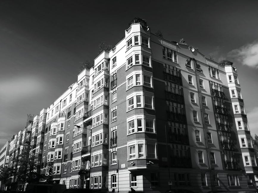 Black and white photograph of a multi-story residential building with numerous windows, balconies, and external fire escape stairs. The building is situated on a paved street with a few trees visible at the base. Shadows cast by the building highlight its architectural features, and the sky appears partly cloudy. This image, associated with house removals and relocation services, illustrates the type of property involved in local flat and apartment moves, as handled by Elephant and Castle removals. The scene captures the urban environment where furniture transport and packing for home relocation are typically conducted, with the building's height and window arrangement emphasizing its residential nature and the logistics involved in moving furniture and belongings in multi-storey buildings.