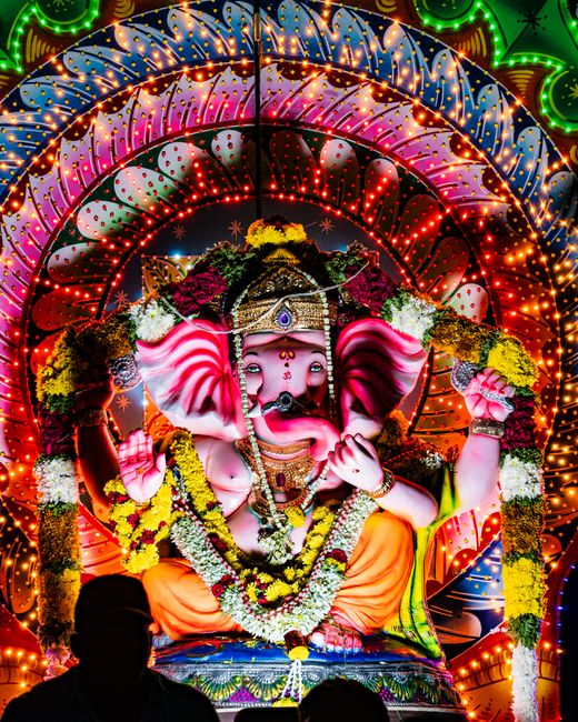 A brightly lit, colorful scene featuring a large illuminated installation of Lord Ganesha, the Hindu deity with an elephant head, seated on a decorated platform. The display is set against a multi-tie
