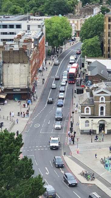 An aerial view of Walworth Road SE17 showing a busy urban street with multiple vehicles including cars and buses moving along the road. Pedestrians are walking on the sidewalks, some near the bus stops, and a cyclist is seen crossing at an intersection. On the left side, there are various commercial buildings, including a shop with a sign that reads 'LA PASTA', with some people entering and exiting. On the right, a historic-looking Victorian-style building and small residential structures are visible, surrounded by green trees lining the street. The scene captures the typical environment of a neighbourhood undergoing home relocation or furniture transport, as part of the packing and moving process, with vehicles likely preparing for or during a local removals service by elephantandcastlemanandvan.com, situated in a central London area.